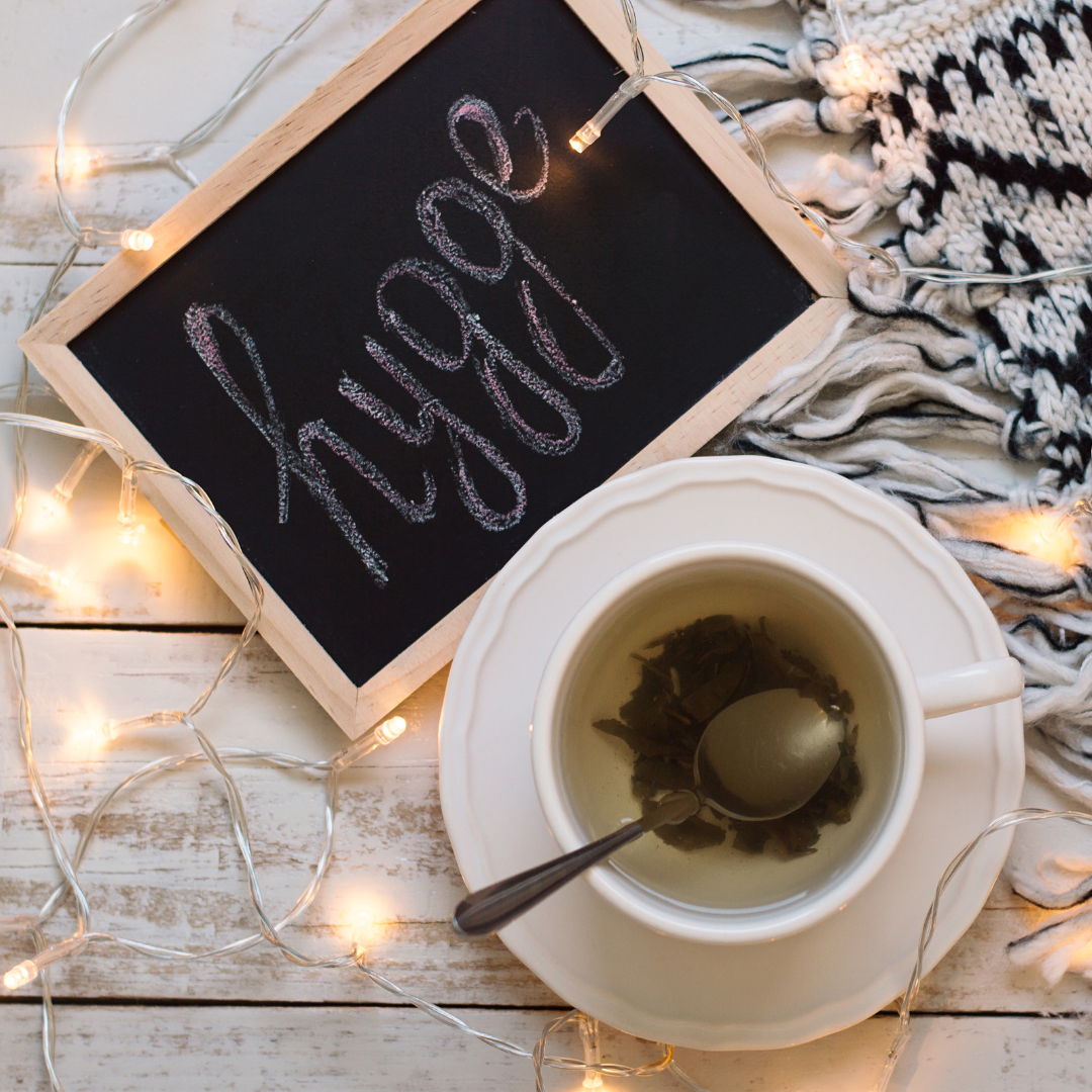 Chalkboard with "hygge," cup of tea, fairy lights, and a cozy knitted blanket.