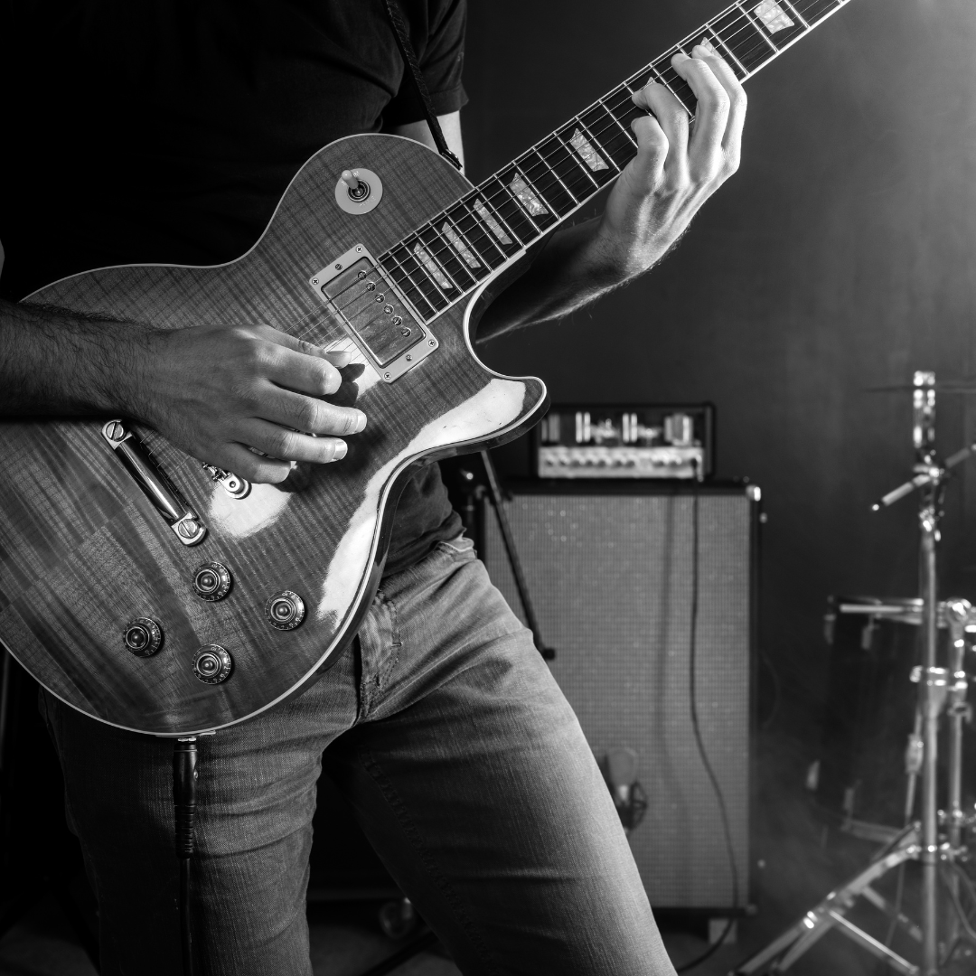 A person playing an electric guitar with an amplifier and drums in the background. Black and white photo.