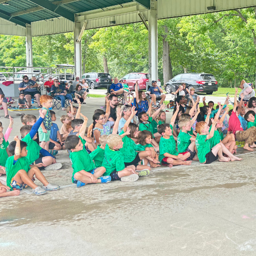 A group of children in green shirts are sitting and raising their hands in a covered outdoor area, while adults observe.