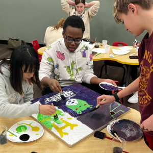 Kids painting on a canvas at a table with paint and brushes, engaged in an art project.