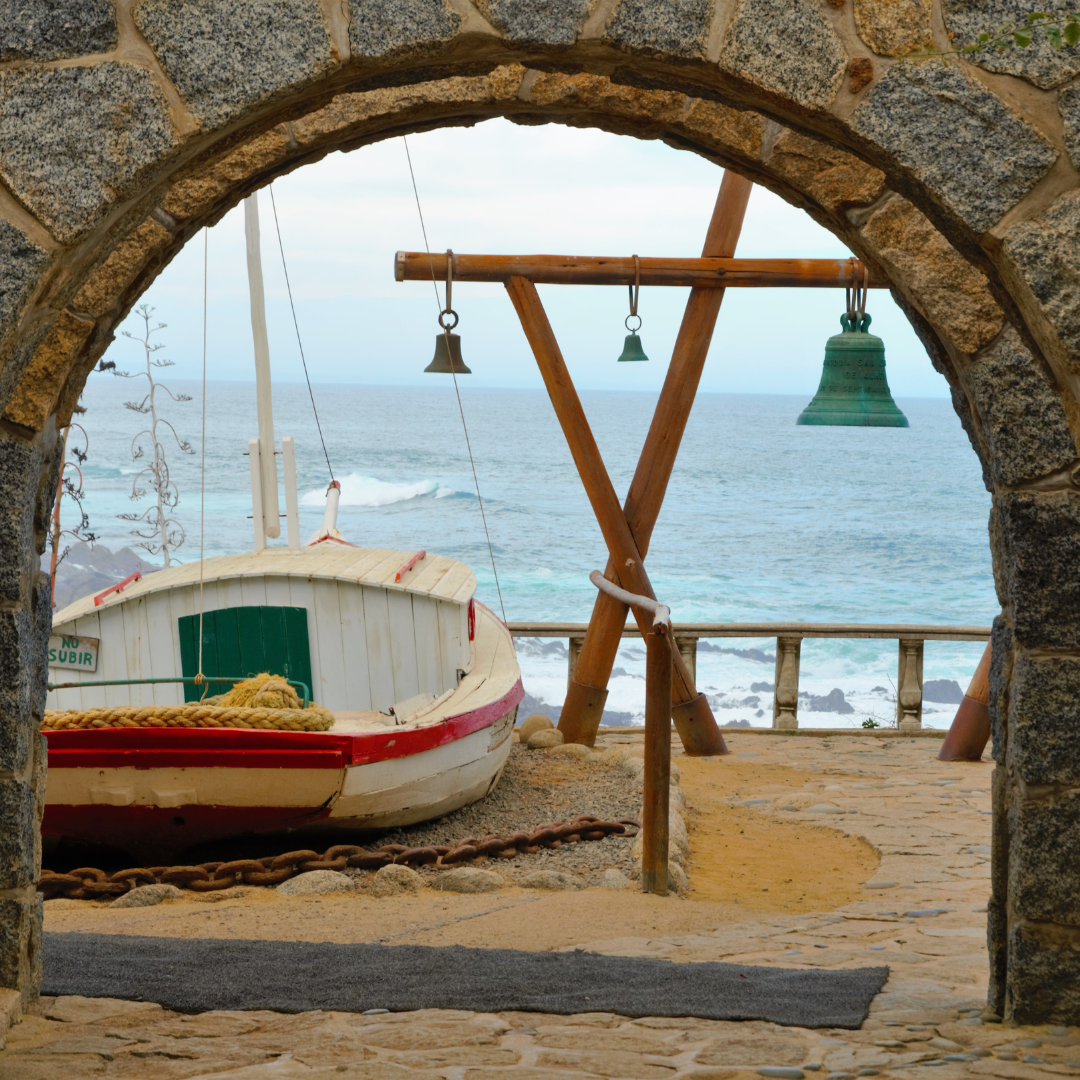A wooden boat and bells viewed through a stone archway by the sea.
