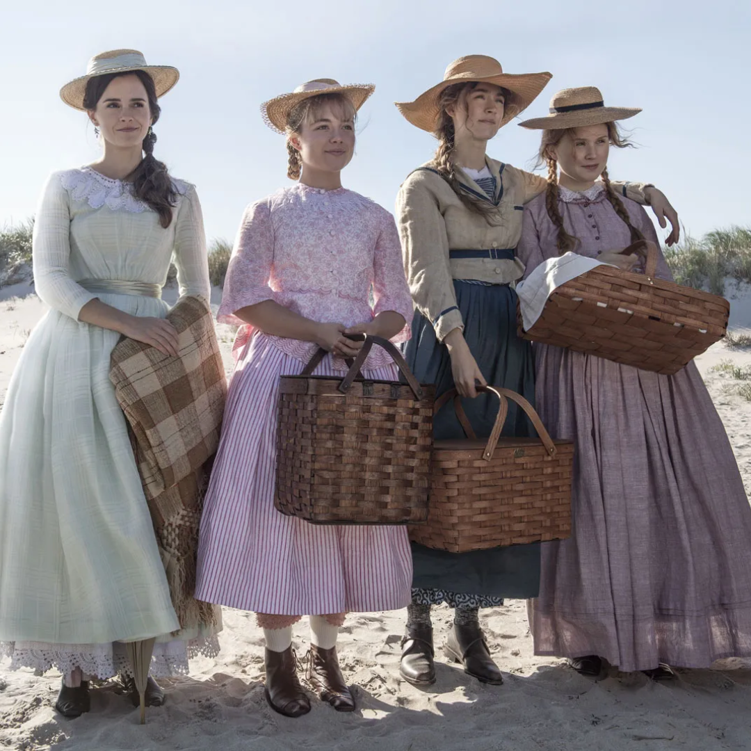 Four women in vintage outfits with straw hats, holding picnic baskets, stand on a sandy beach.