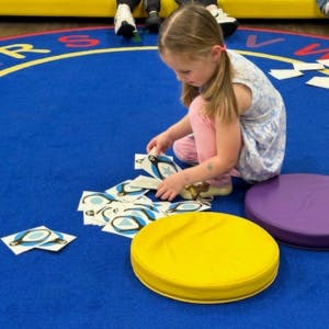 A child playing with cards on a circular alphabet rug near colorful cushions.