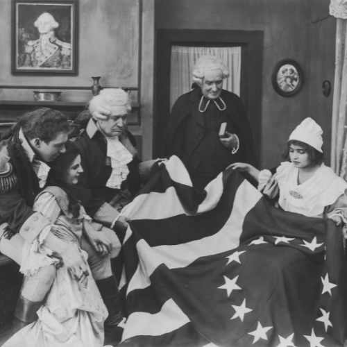 A historical scene showing figures in 18th-century attire, gathered around a woman holding an American flag.