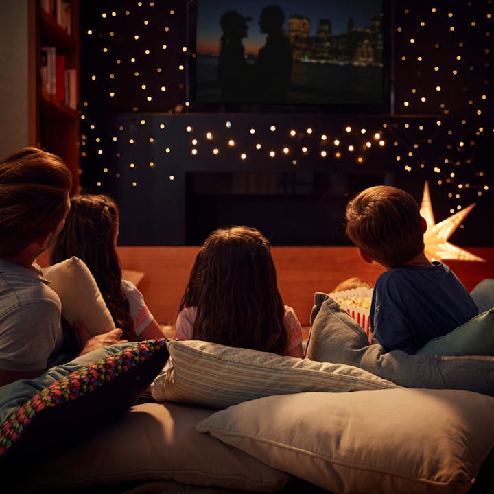A cozy movie night with kids watching TV, fairy lights, pillows, and popcorn.