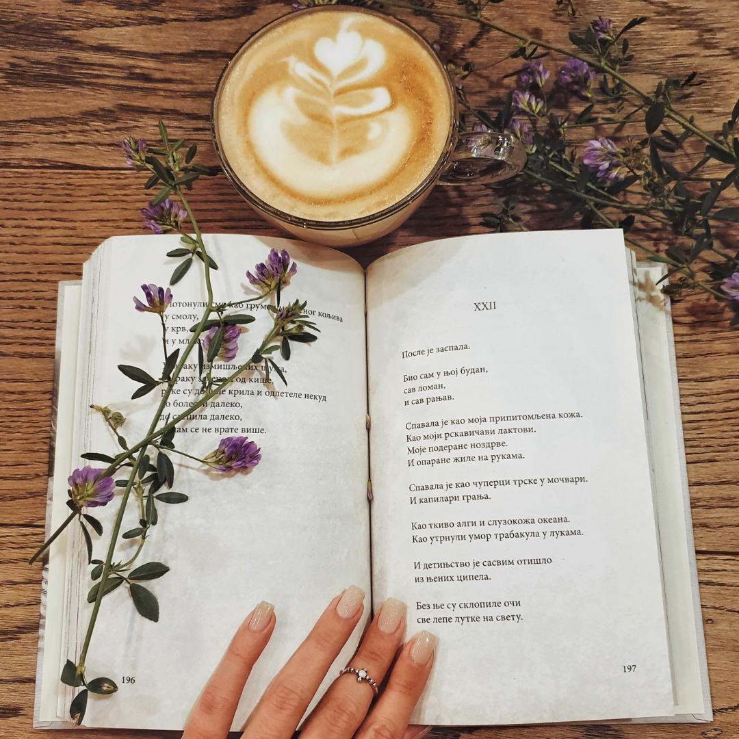 Open book with flowers, a latte with latte art, and a hand with a ring on a wooden table.