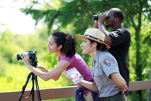 Three people are outdoors: two are taking pictures with cameras, while one, wearing a ranger hat, holds a booklet and observes.