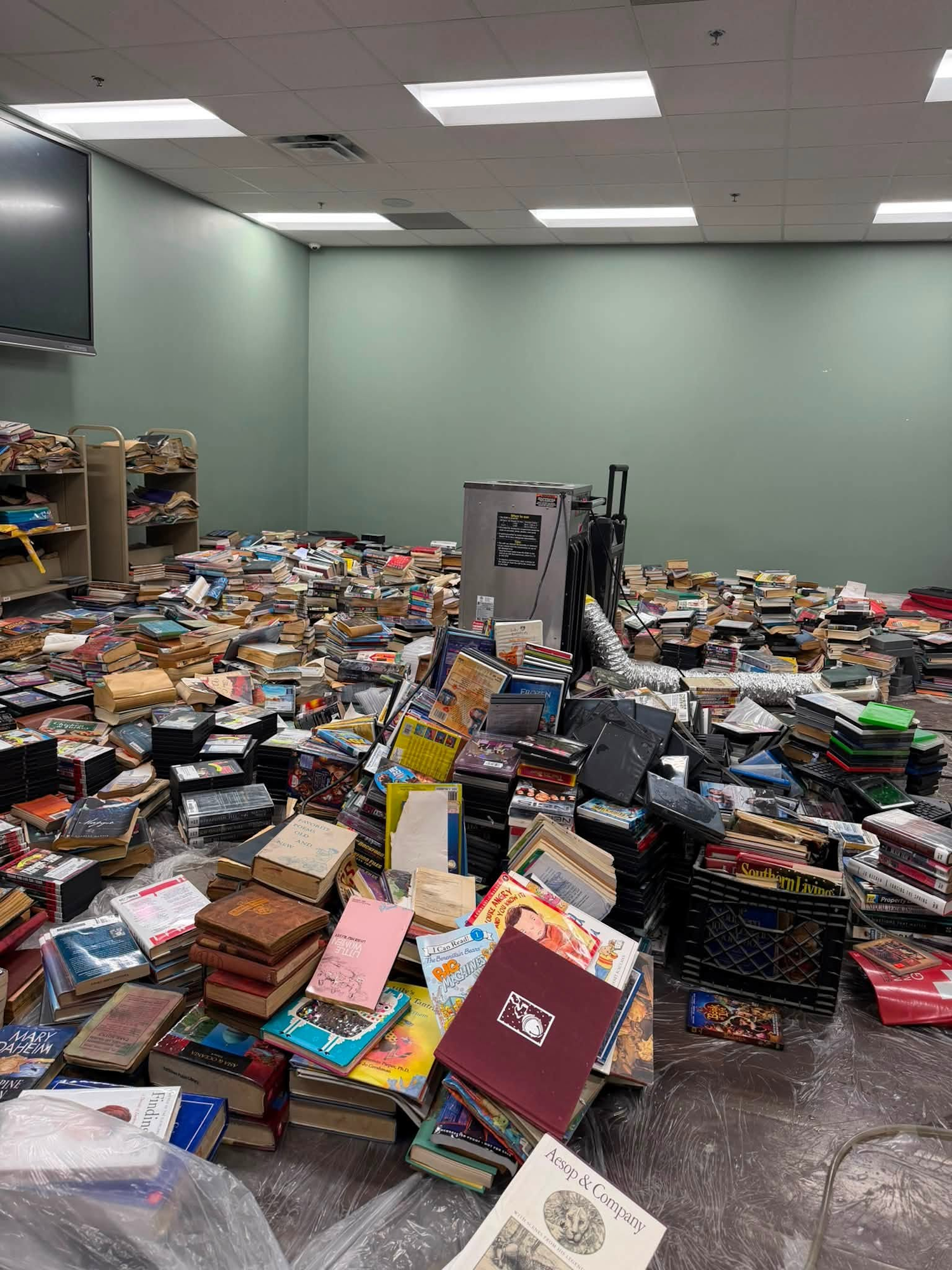 A chaotic room filled with piles of books and media, scattered across the floor, with some shelves in the background.