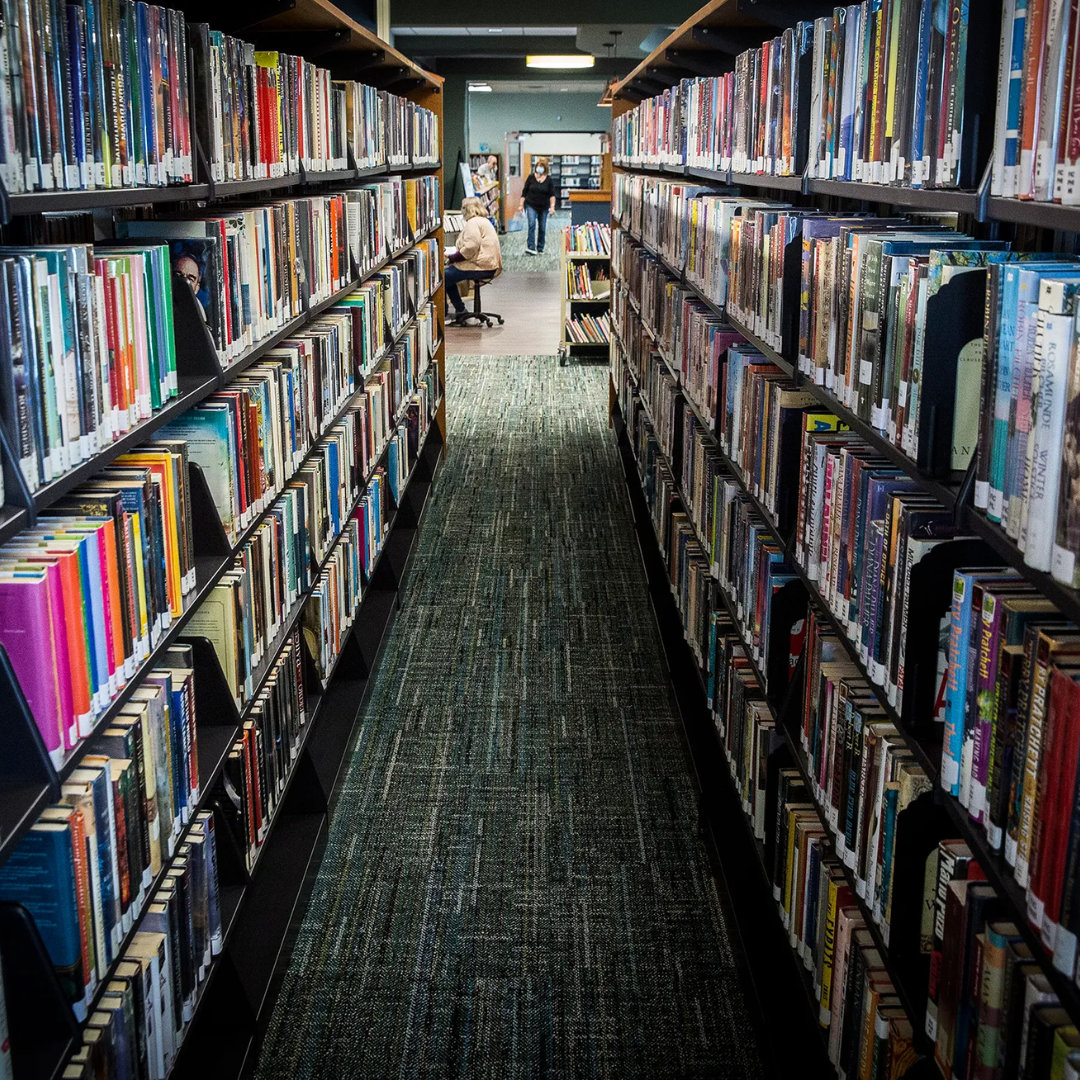 A library aisle with bookshelves full of colorful books, leading to a seating area with people at tables.
