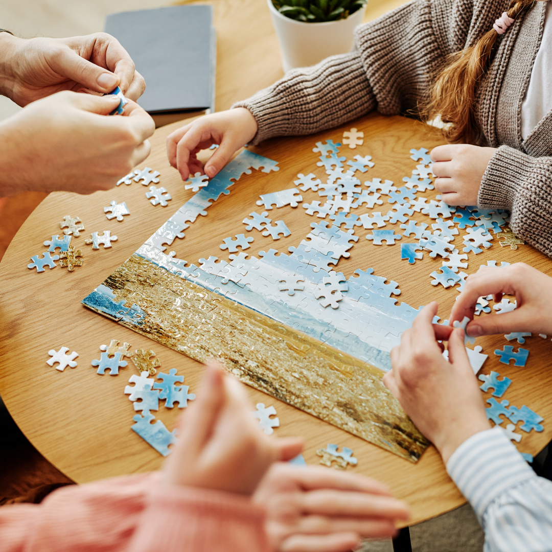 Four people assembling a puzzle with a sky and landscape image on a wooden table.
