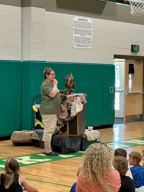 Person speaking indoors, with an owl perched on a stand, surrounded by an audience.