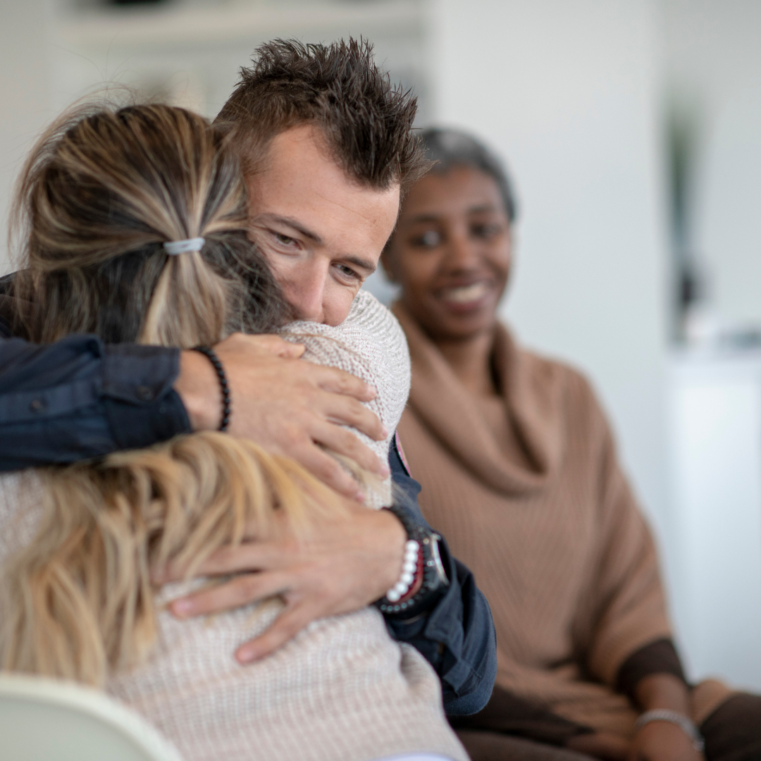 Two people are hugging while another person watches and smiles.