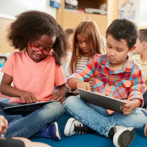 Children sitting on the floor, using a tablet and reading books in a classroom setting.
