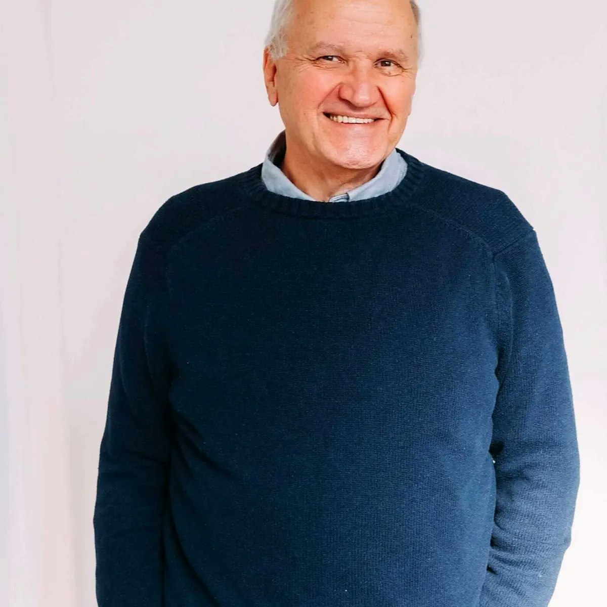 A smiling older man wearing a navy sweater stands against a light background.