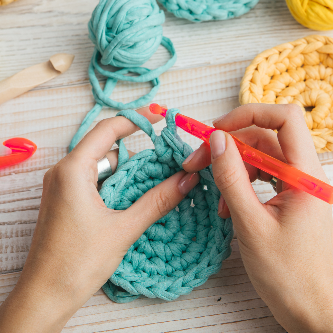Hands crocheting with chunky teal yarn, red crochet hook, more yarn balls and a wooden hook nearby.