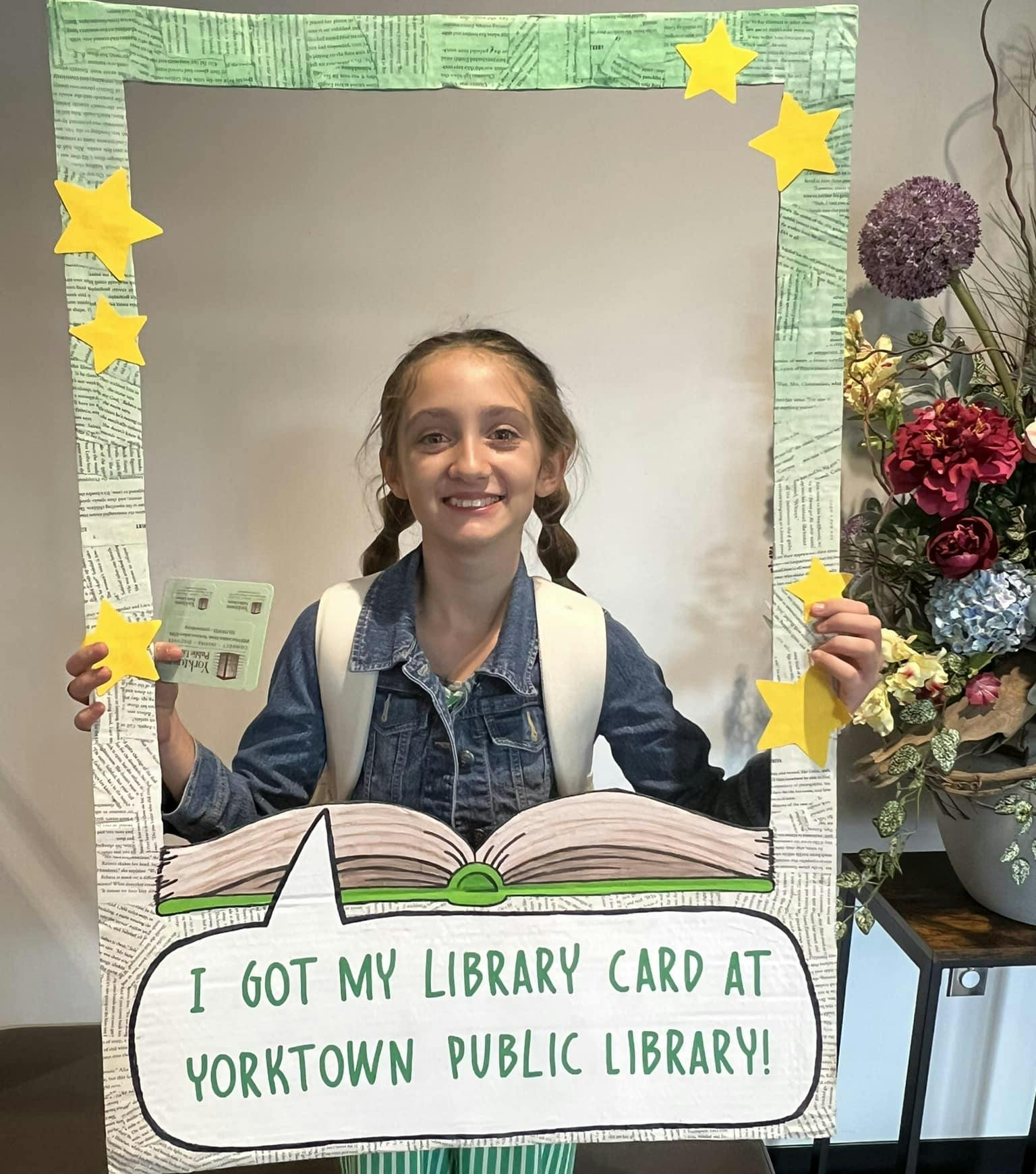 A smiling girl holding a library card, standing behind a frame saying "I got my library card at Yorktown Public Library!"