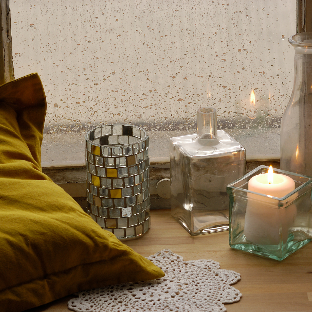 Cozy scene with a yellow pillow, a candle in a glass holder, a mirrored vase, and a crocheted doily near a rainy window.