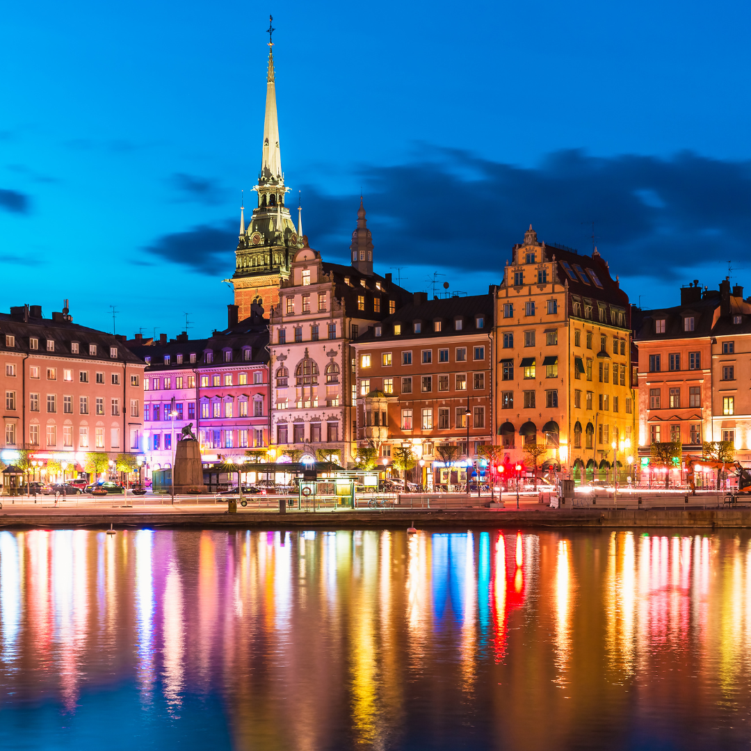 A waterfront view of colorful buildings and a church spire at night, with reflections on the water.