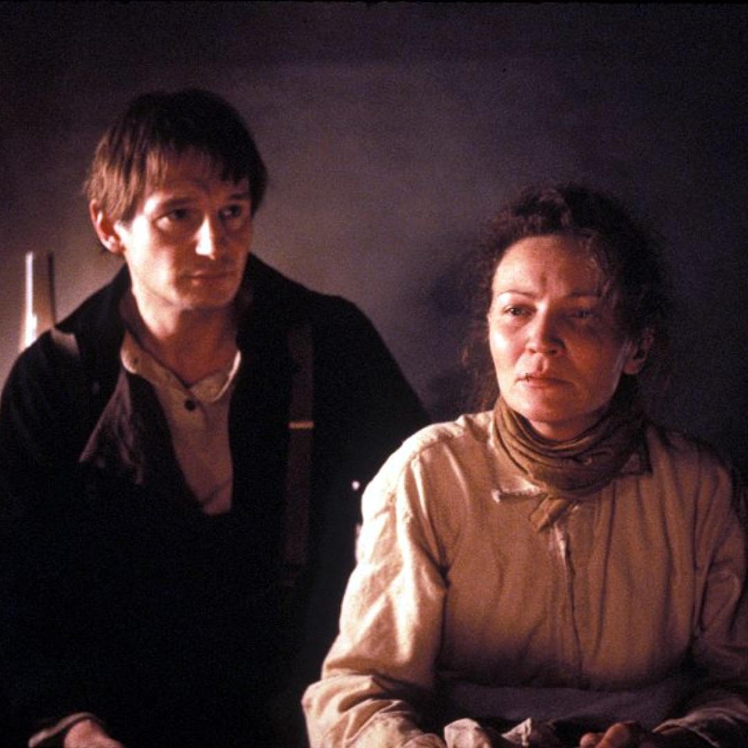 Two people in historical attire sit side by side in a dimly lit setting.