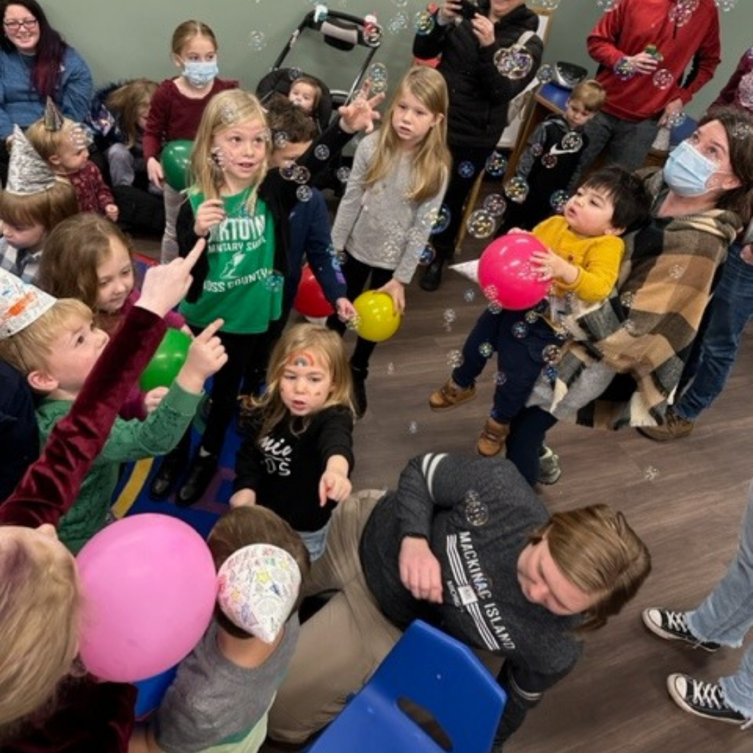 Children and adults at a party with balloons and bubbles.