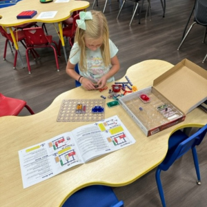 A child playing with educational toys at a classroom table, with instruction sheets and colorful elements visible.