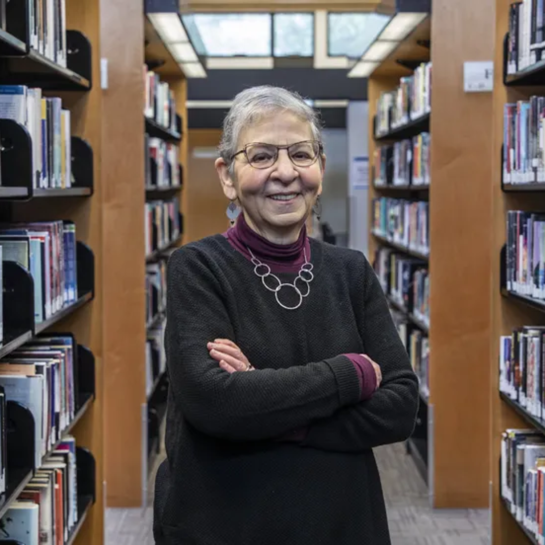 A person with glasses stands smiling in a library aisle surrounded by bookshelves.