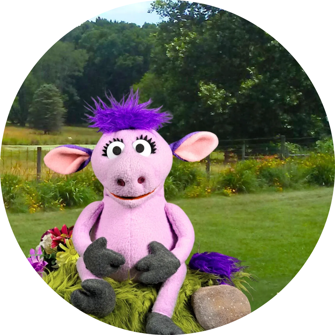 A purple, plush-like creature with purple hair, large eyes, and big ears, sitting outdoors with greenery and a field in the background.