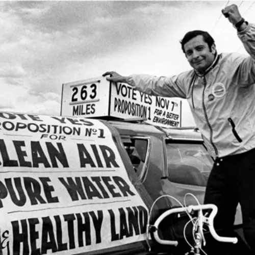 A man promotes a campaign for clean air, pure water, and healthy land, standing next to a car with signs for voting on environmental issues.