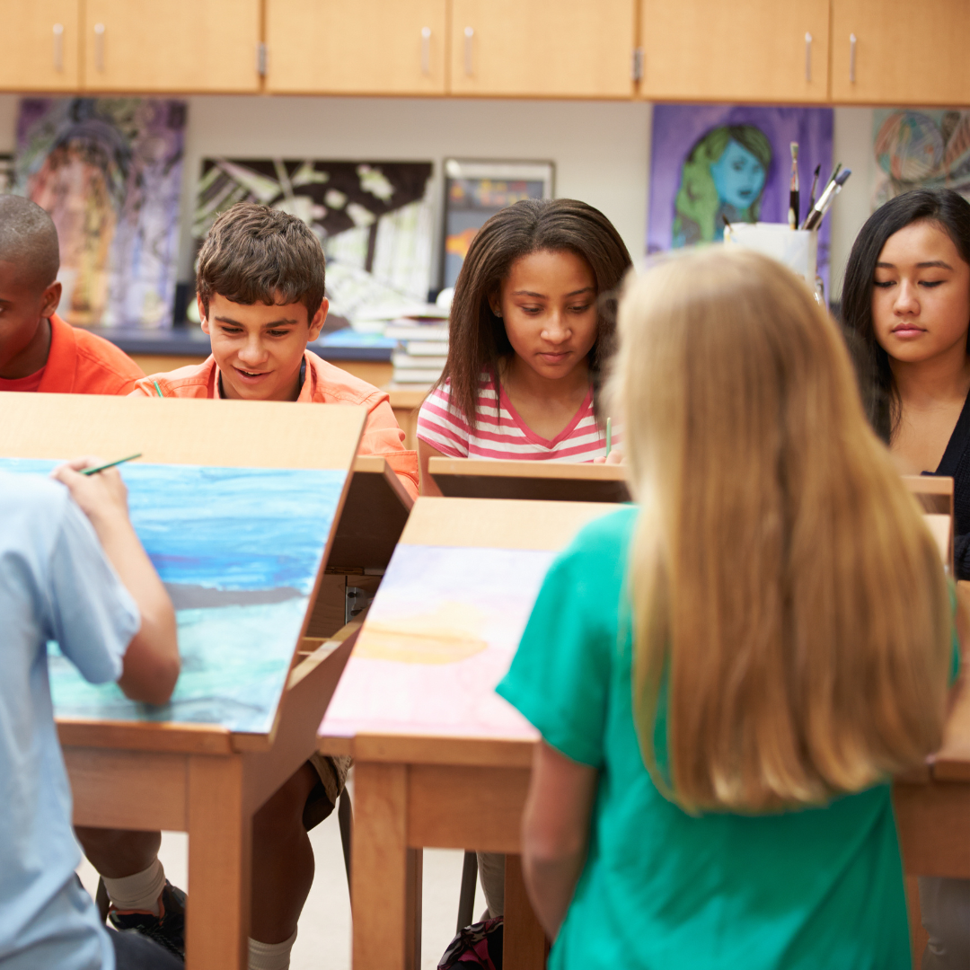 Students painting in an art class, sitting at easels, with art supplies and paintings in the background.
