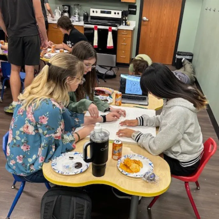 People gathered around a table with snacks, drinks, and an open book, in a room with kitchen appliances and a laptop.