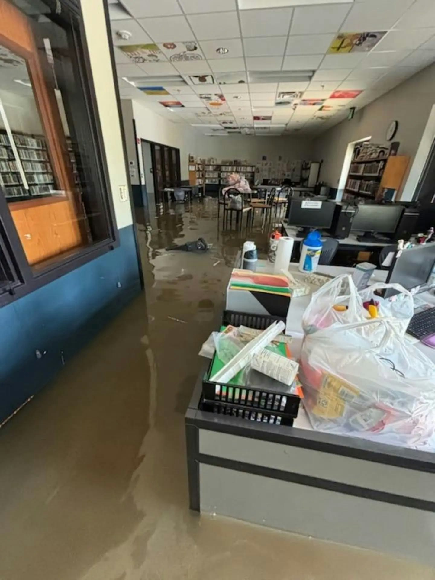 The image shows a flooded room with water covering the floor, desks, and scattered items, indicating a significant water damage event.