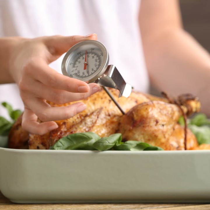 A person is using a meat thermometer to check the temperature of a roasted chicken on a bed of basil leaves.