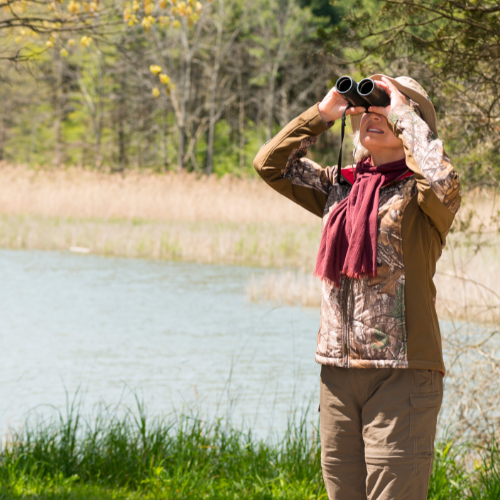 A person is outdoors by a lake, using binoculars and wearing a hat and scarf, enjoying nature and birdwatching.