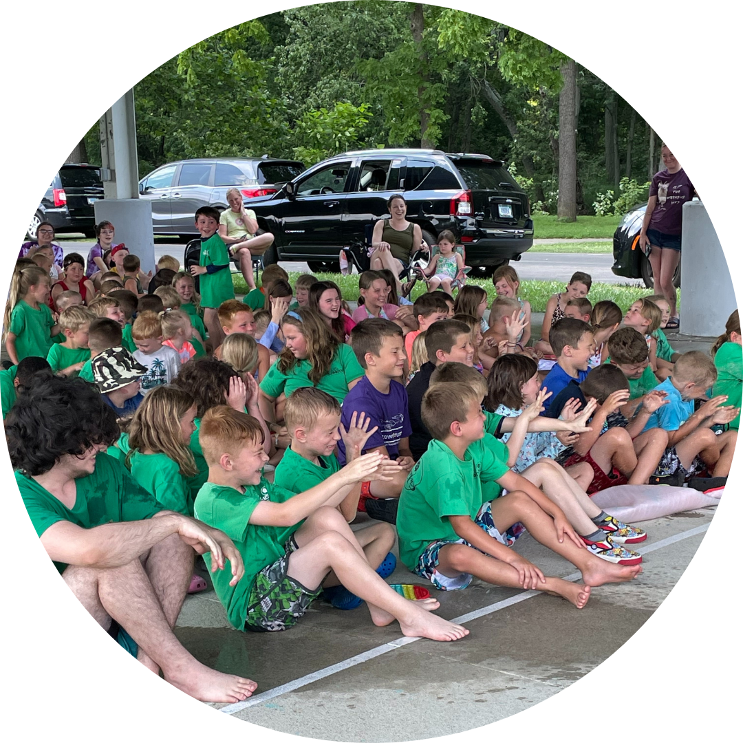 Group of children sitting together, some clapping, wearing green shirts, with adults present, likely at an event or gathering.