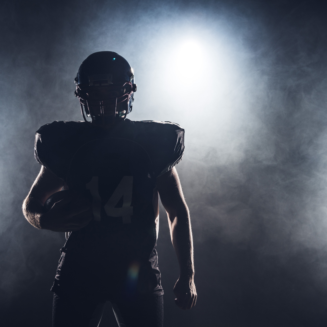 Football player in helmet and jersey with number 14, holding a ball, backlit and surrounded by haze.