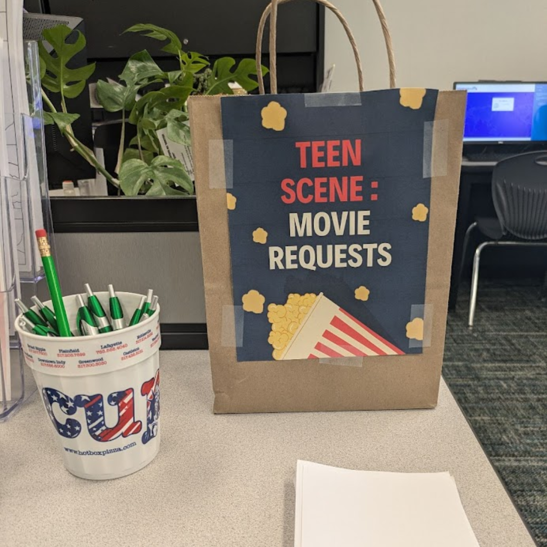 A paper bag labeled "TEEN SCENE: MOVIE REQUESTS," a cup with pens, a plant, and a computer in the background.