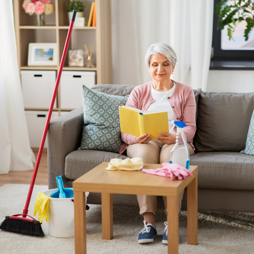 A woman sits on a sofa reading a book, surrounded by cleaning supplies on a table.