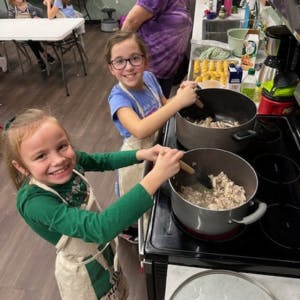 Two smiling children cooking, stirring pots on a stove in a kitchen.