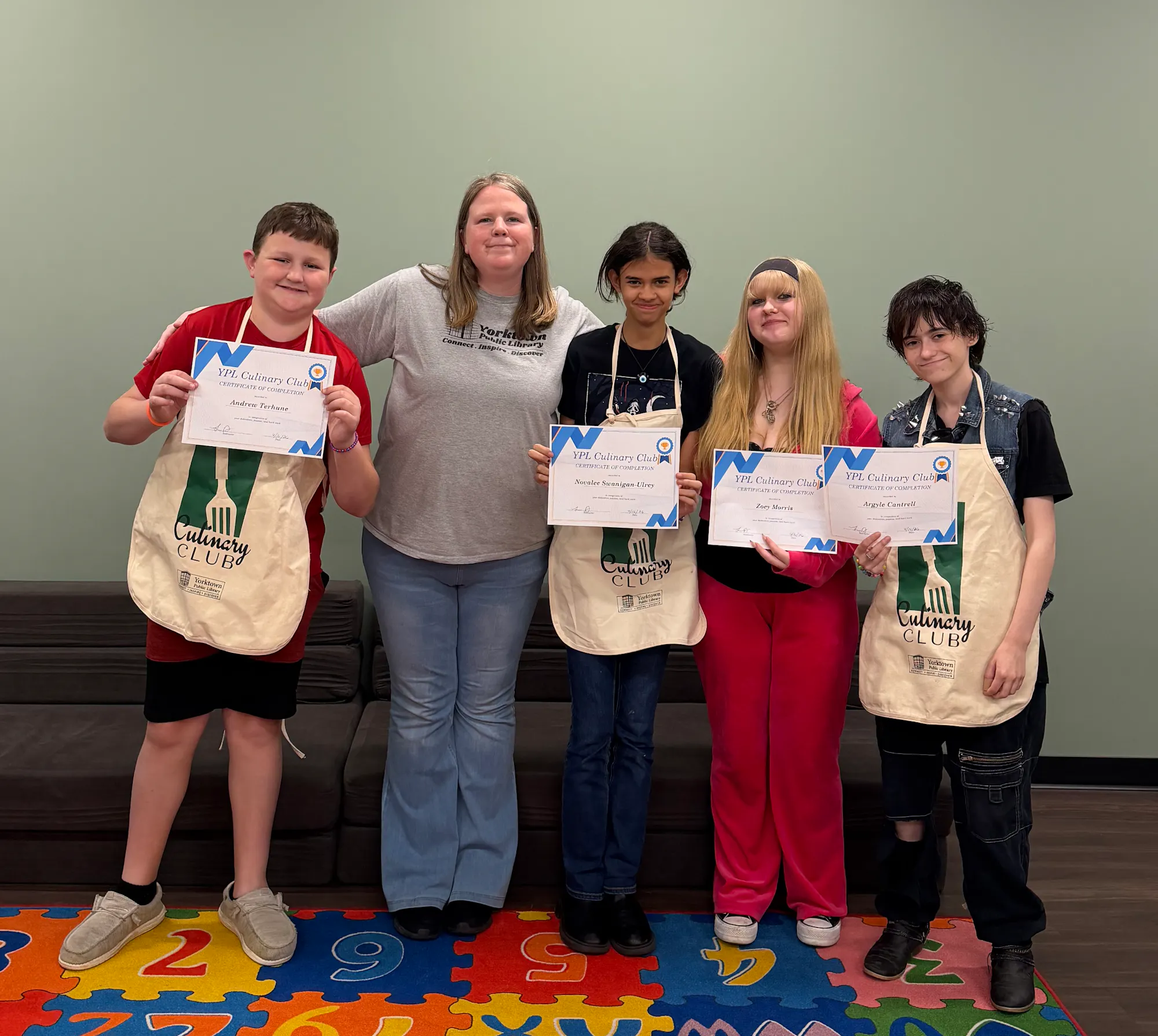 A group of five individuals holds up certificates in an indoor space with a colorful alphabet mat.