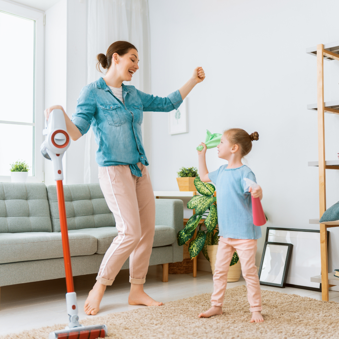 A woman and a child joyfully cleaning a living room. The woman is holding a vacuum, and the child has cleaning supplies.