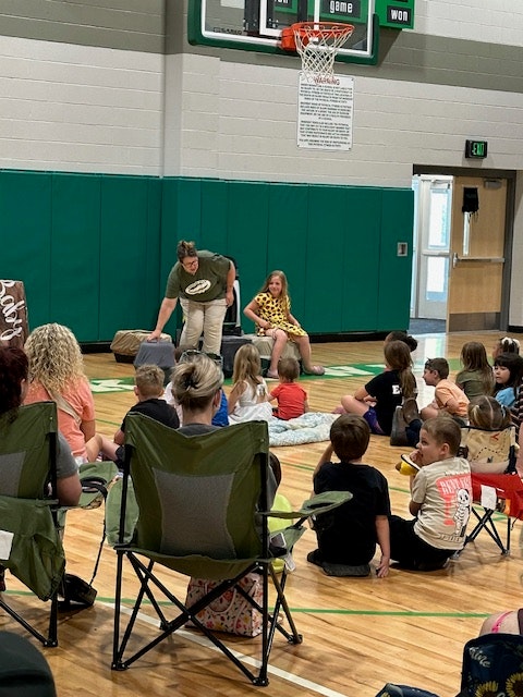 A group of people, including children, seated on a gym floor, listening to two people standing in front near a basketball hoop.
