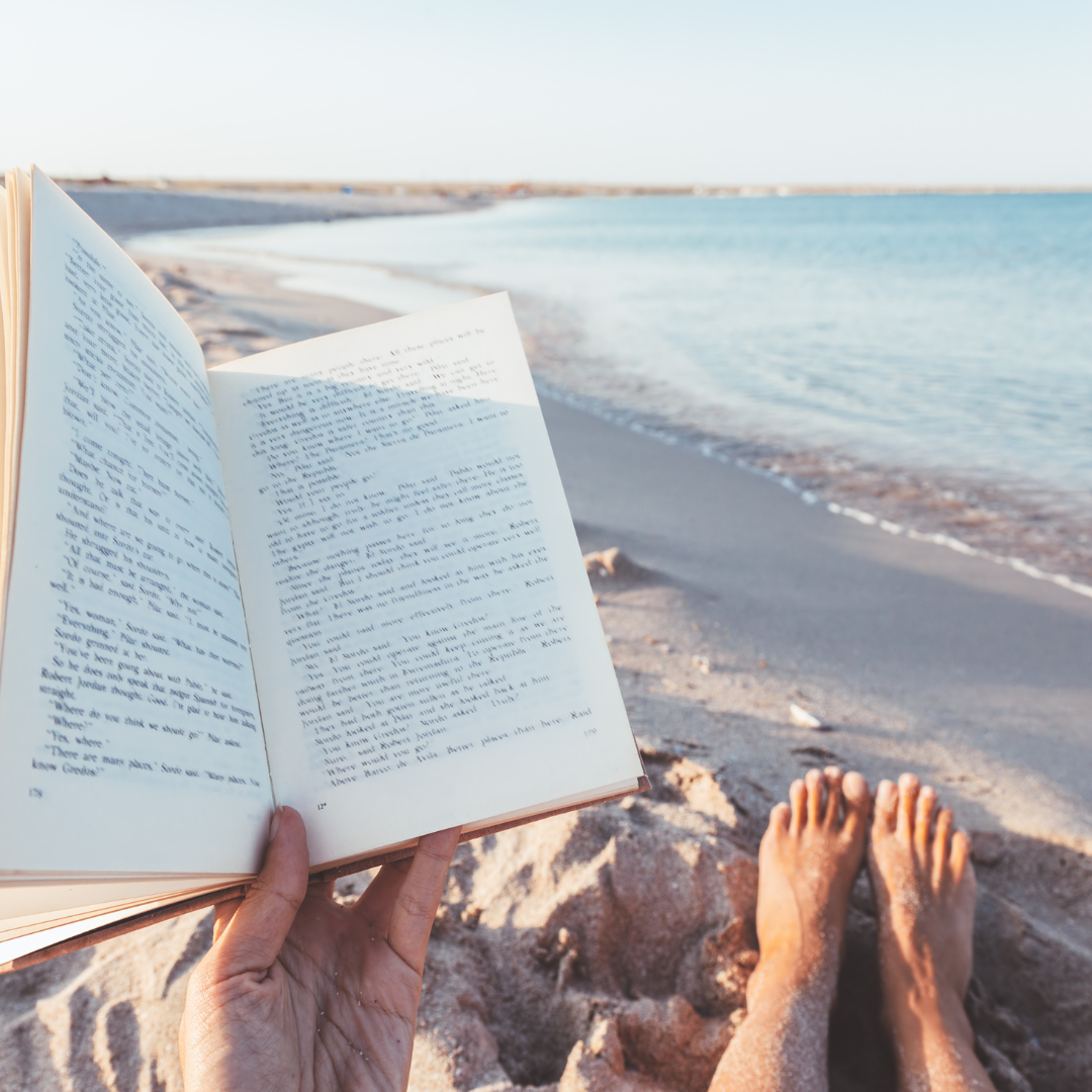 Person reading a book on a sandy beach with the ocean in the background.
