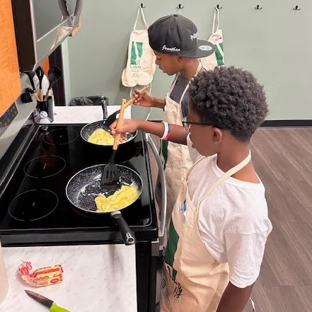 Two kids are cooking eggs on a stovetop, wearing aprons, and focused on preparing their meal.