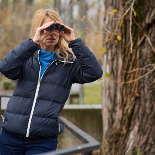 A woman in a dark jacket uses binoculars outdoors, observing her surroundings near a tree and a railing.