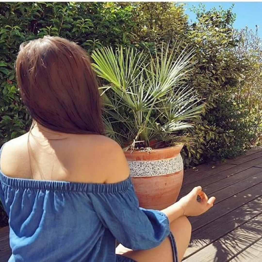 A person sitting on a wooden deck in a blue off-shoulder top, meditating near a potted plant, surrounded by lush greenery.