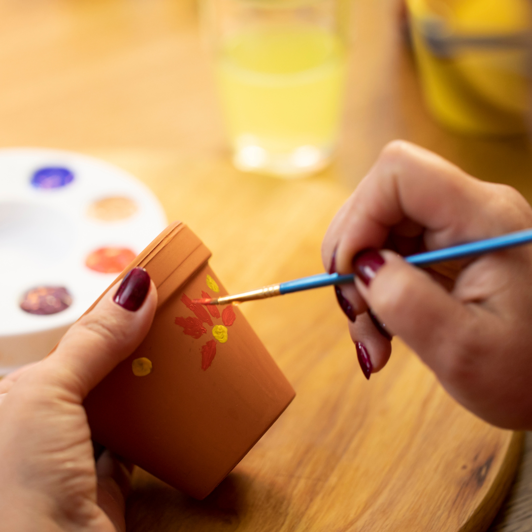 A person is painting a small terracotta pot with colorful designs while holding a paintbrush.