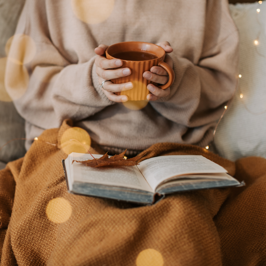 Person holding a cup, open book on lap, warm lighting, cozy atmosphere.