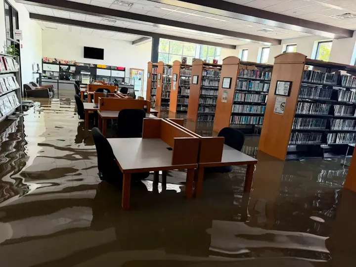 The image shows a library with water flooding the floor, submerging tables and chairs, creating a significant mess.