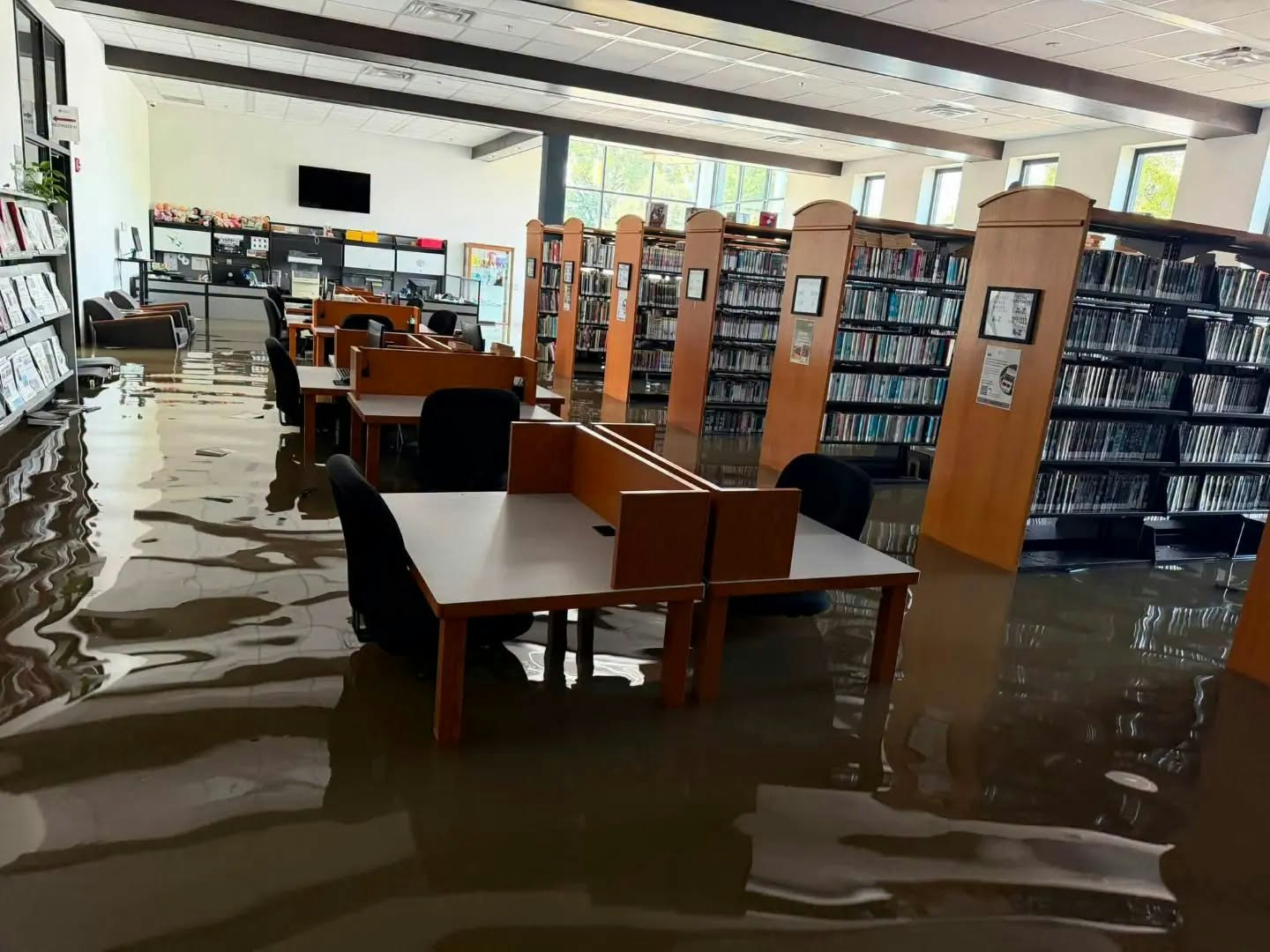 The image shows a library with water flooding the floor, submerging tables and chairs, creating a significant mess.