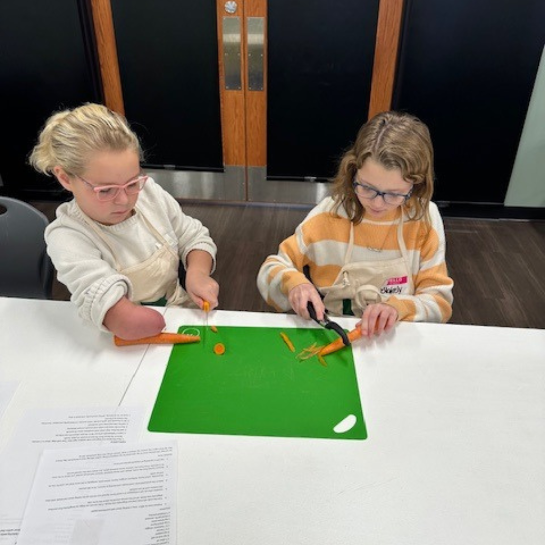 Two children are cutting carrots on a green chopping board at a table.